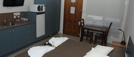 A compact hotel room with a kitchenette, a bed with pillows, a small dining table, and a TV.