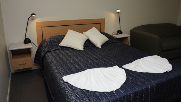 Ein ordentlich bezogenes Bett mit Kopfteil, zwei Nachttische mit Lampen, ein Beistelltisch und ein dunkler Sessel.