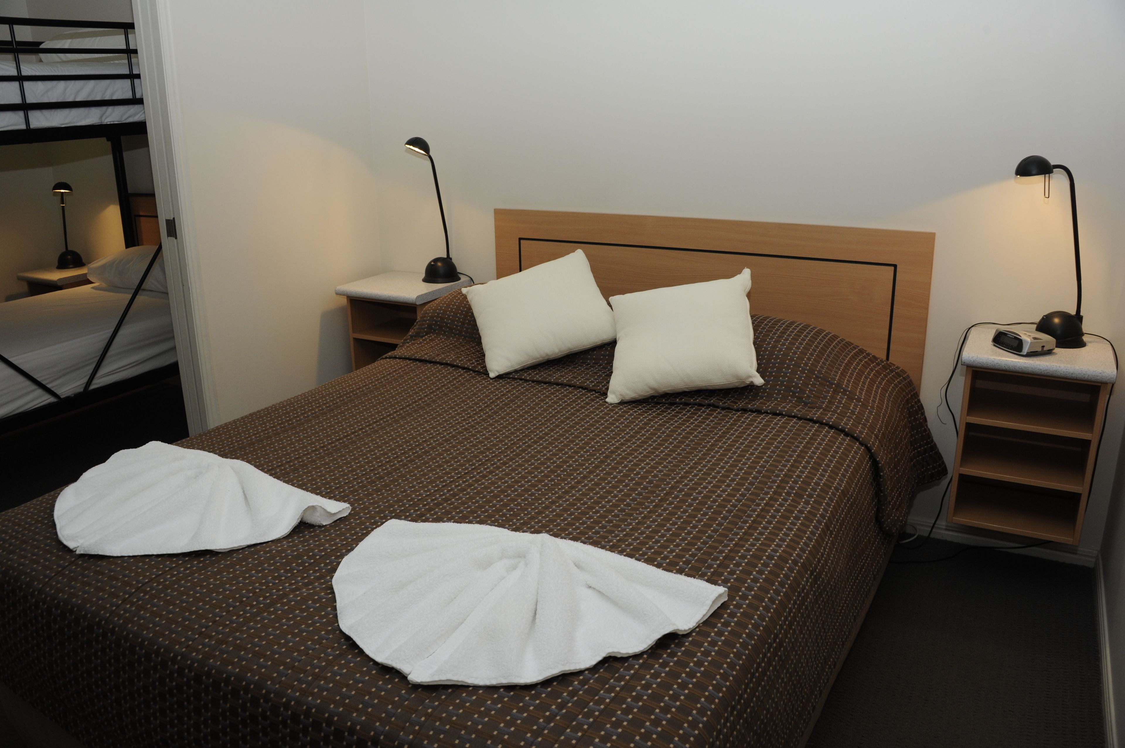 Un lit bien fait, recouvert d’une courtepointe marron à motifs, avec deux oreillers blancs et deux serviettes blanches. Il y a deux tables de chevet avec des lampes, une de chaque côté du lit.