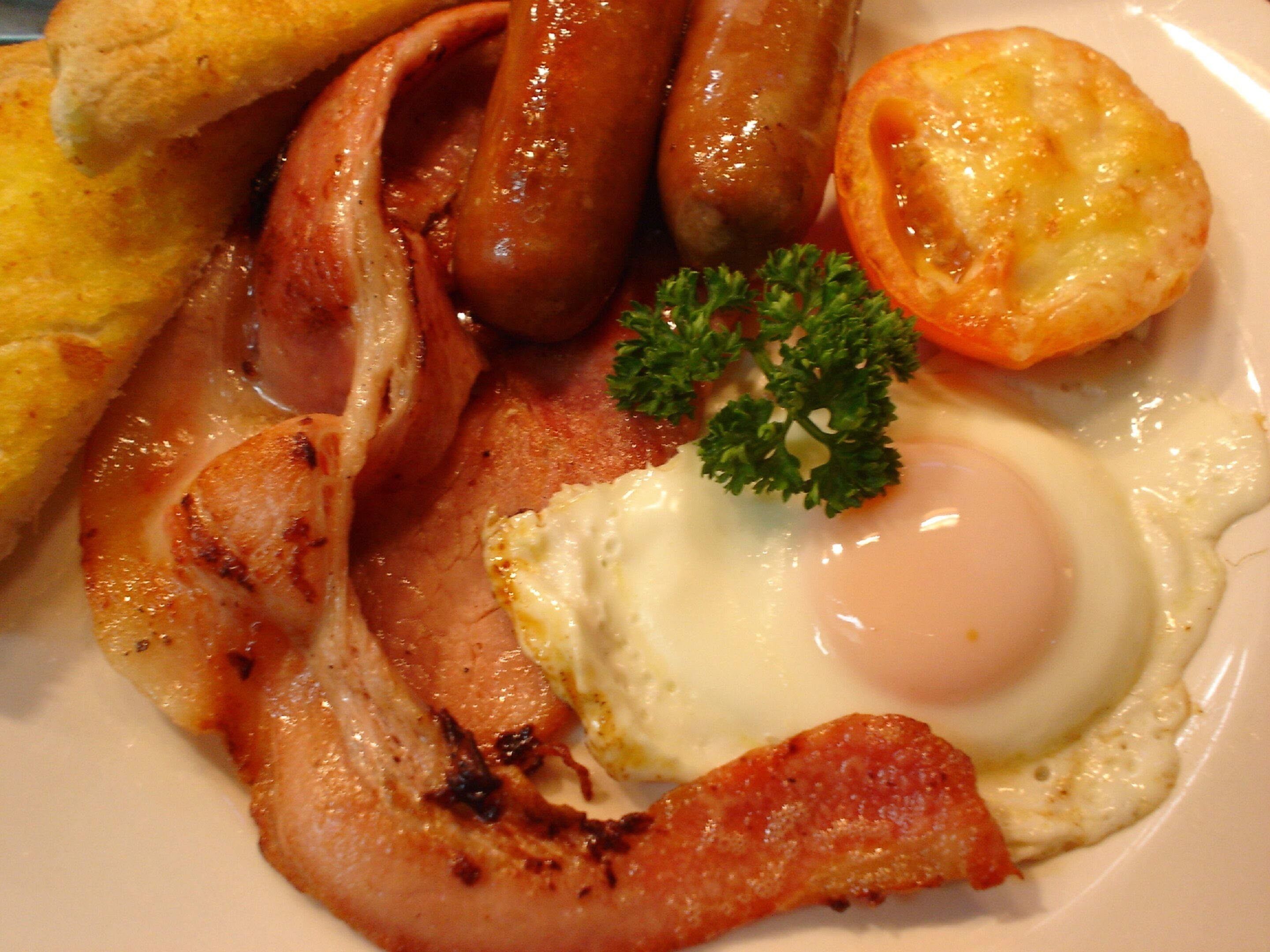A plate of breakfast food including sausages, bacon, a fried egg, toast, and a tomato.