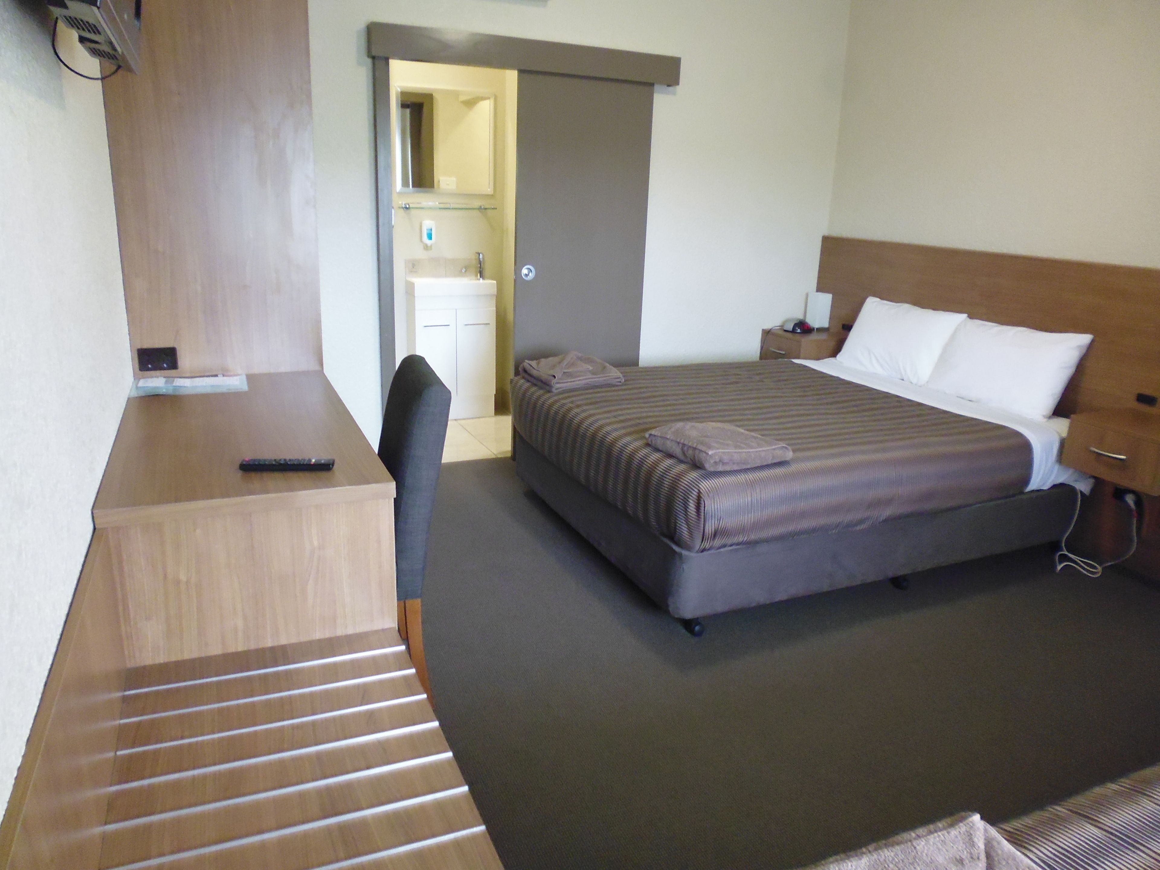 A hotel room with a bed, a desk, a chair, a nightstand, and a bathroom visible through an open door.