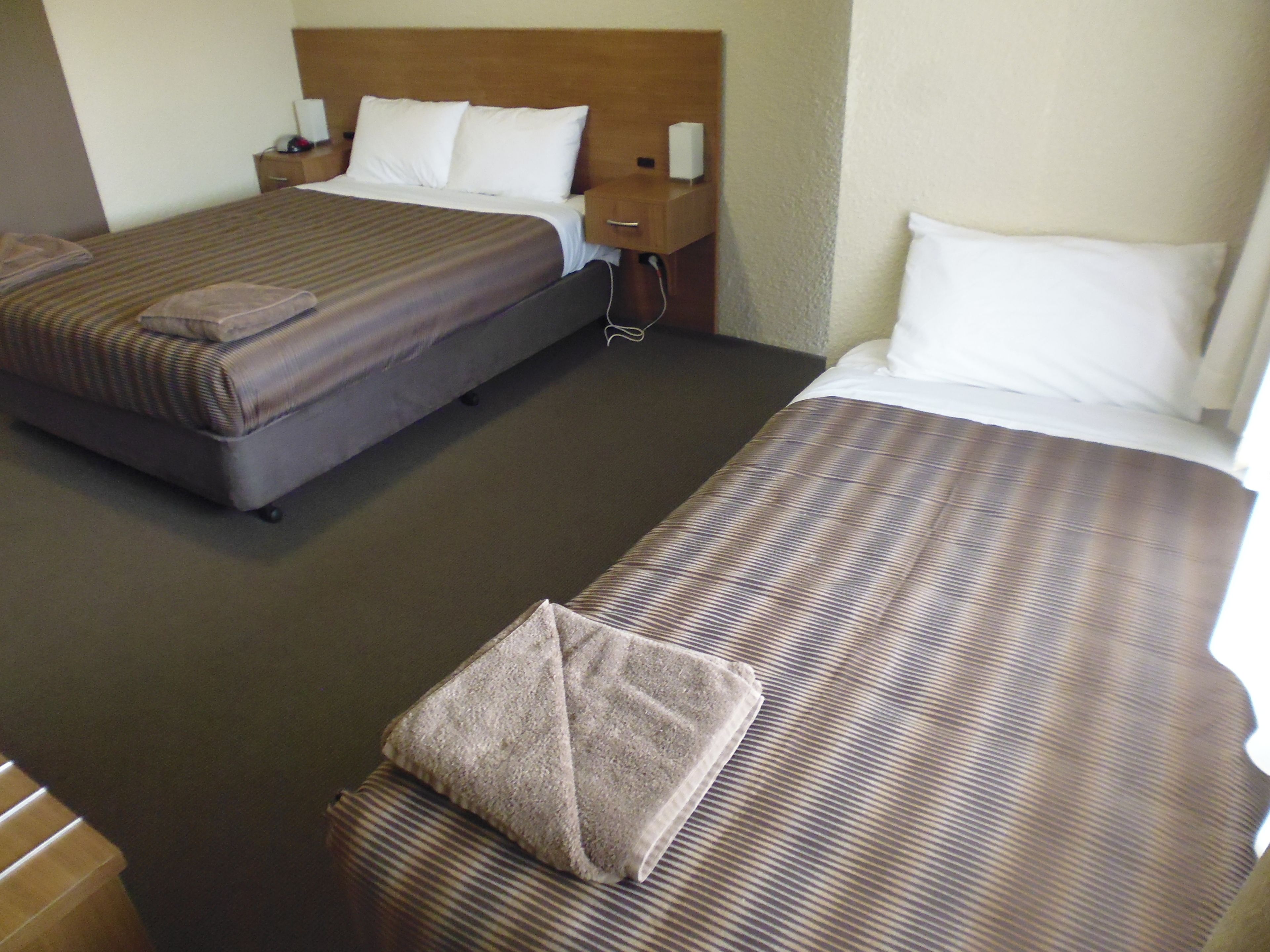 A hotel room with two beds, each with a pillow and a blanket. There is a nightstand with a lamp and a phone on one side.