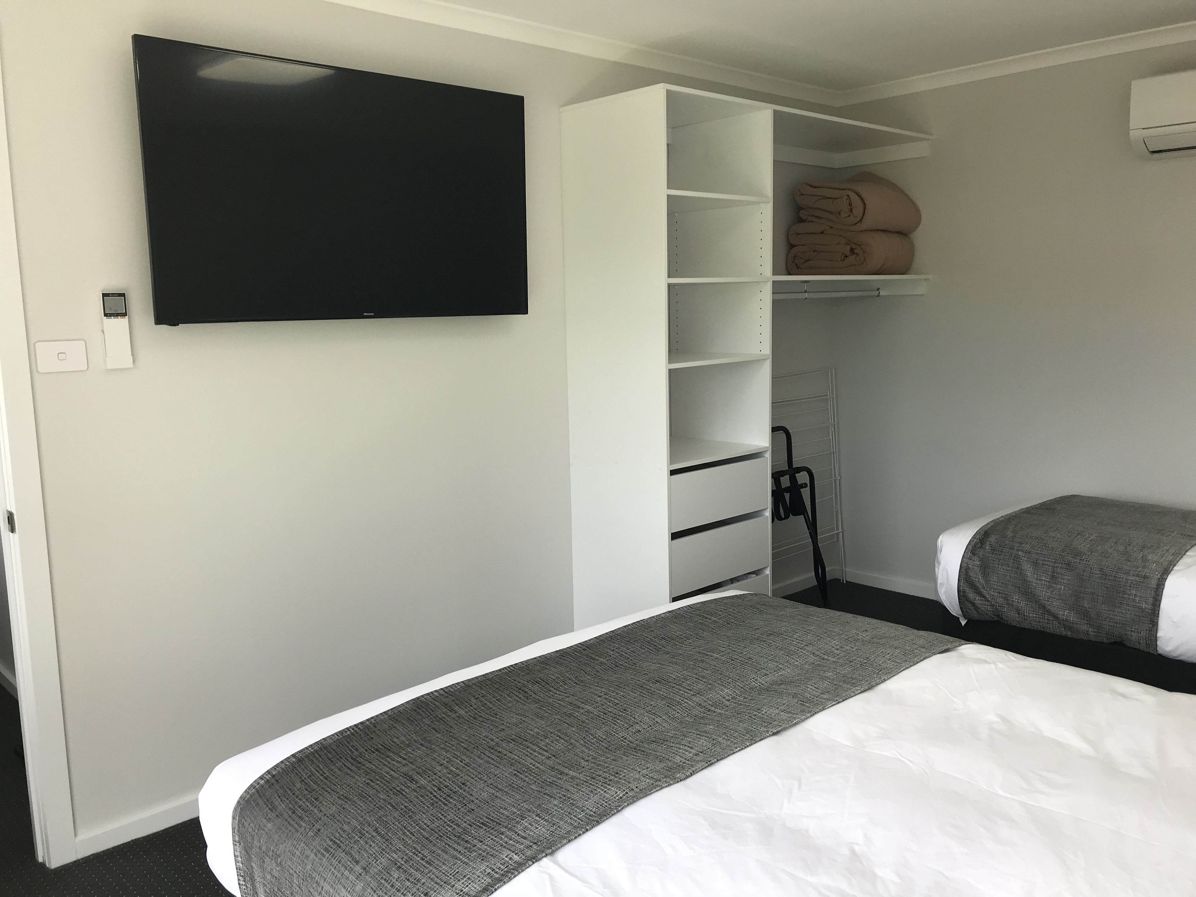 Habitación de hotel con cama, televisor montado en la pared, armario con repisas y una manta doblada sobre la cama.