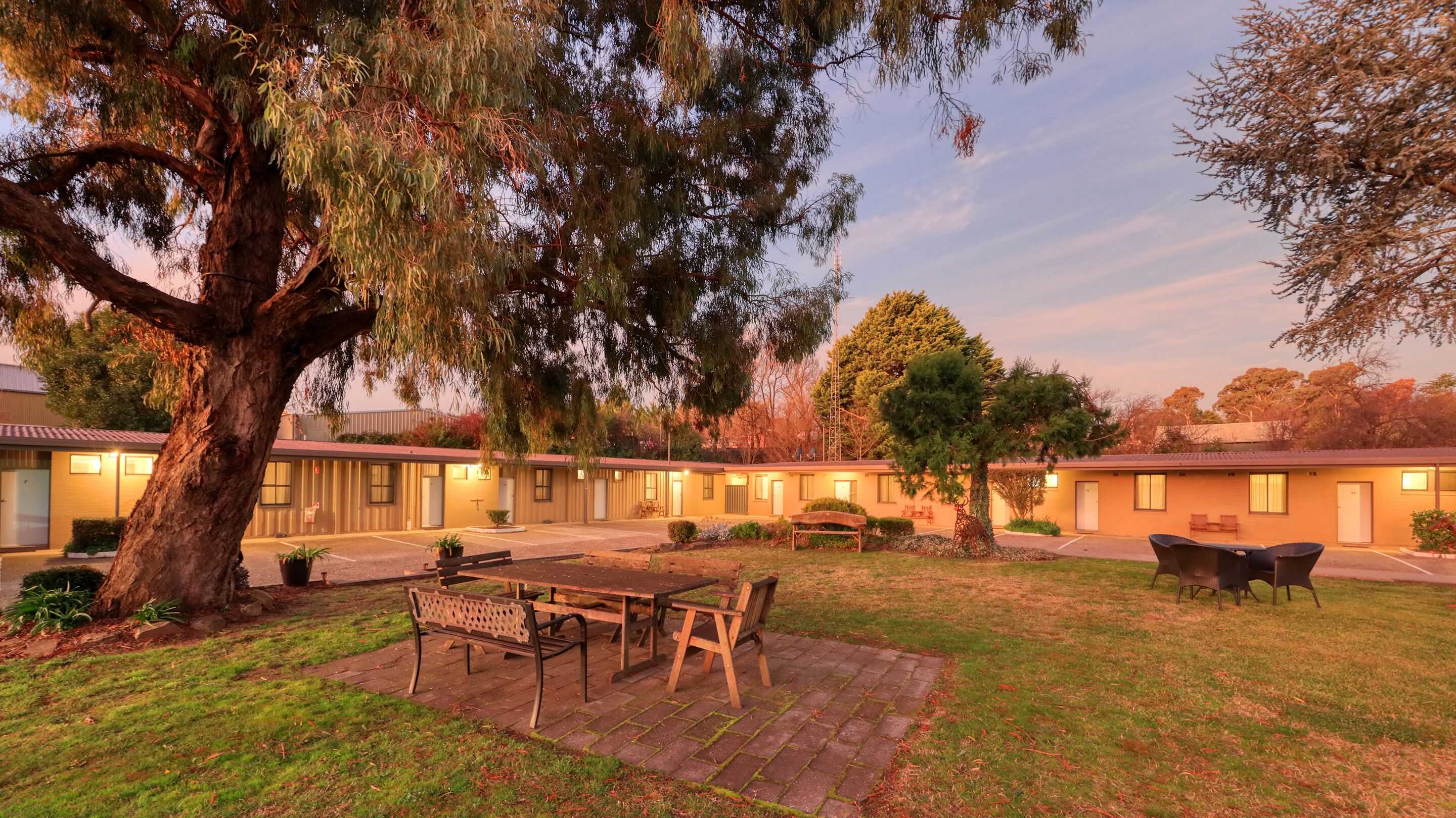 A motel with a large tree in the foreground, a patio area with a table and benches, and a parking lot.