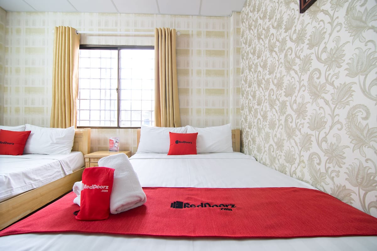 Quarto de hotel com duas camas, cada uma com roupa de cama branca e almofadas vermelhas. Uma toalha vermelha e outra toalha com o logotipo do hotel estão no chão. O quarto tem papel de parede estampado e uma janela com cortinas.