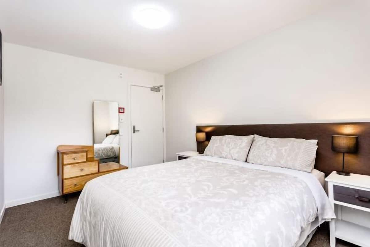 Una habitación de hotel con cama, mesita de noche, lámpara, cómoda y una puerta.