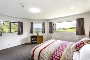 A bedroom with a large bed, a desk, and two windows offering a view of the outdoors.