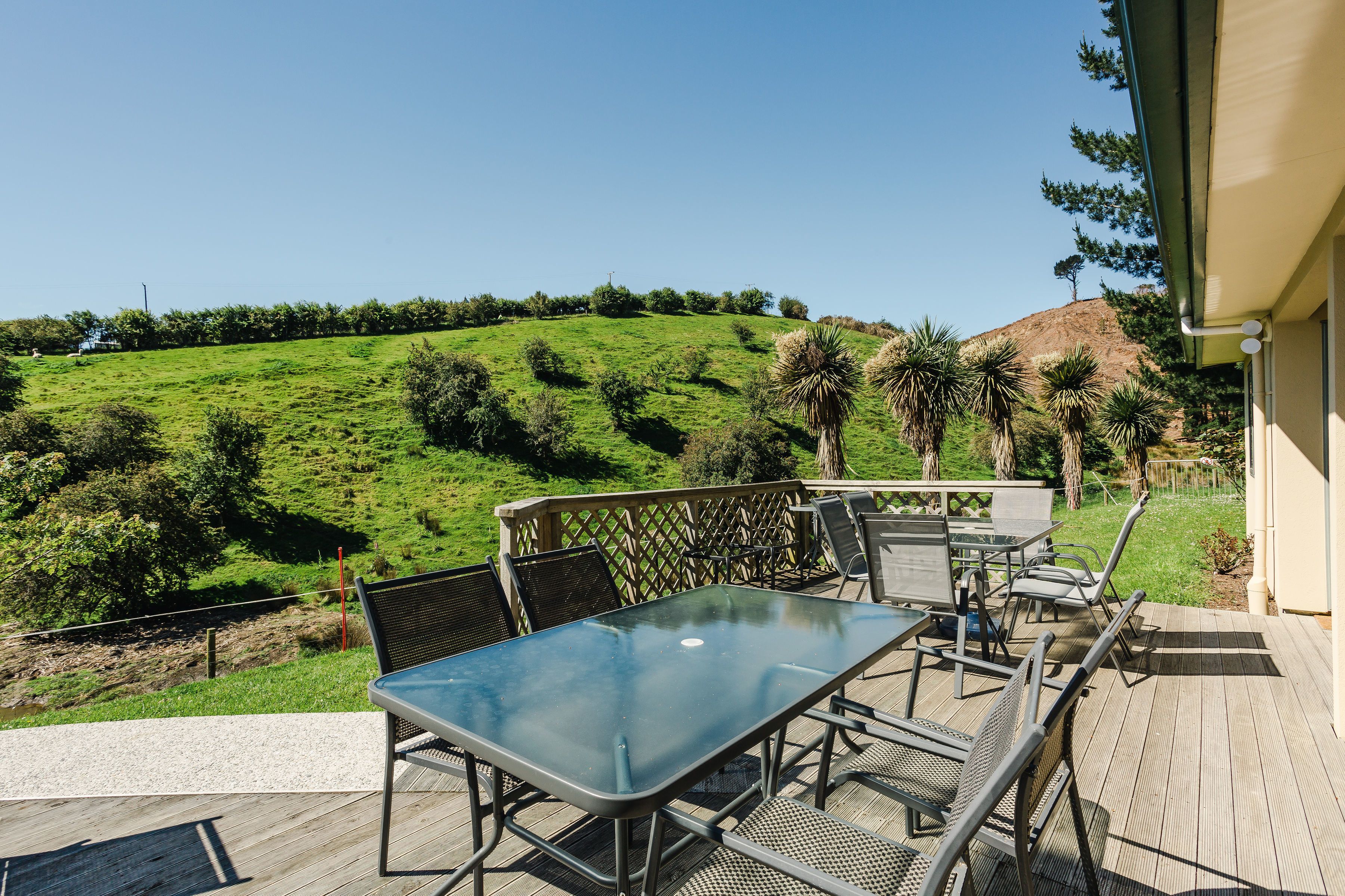 A deck with a glass table and chairs, overlooking a green hillside with trees and a hill in the distance.