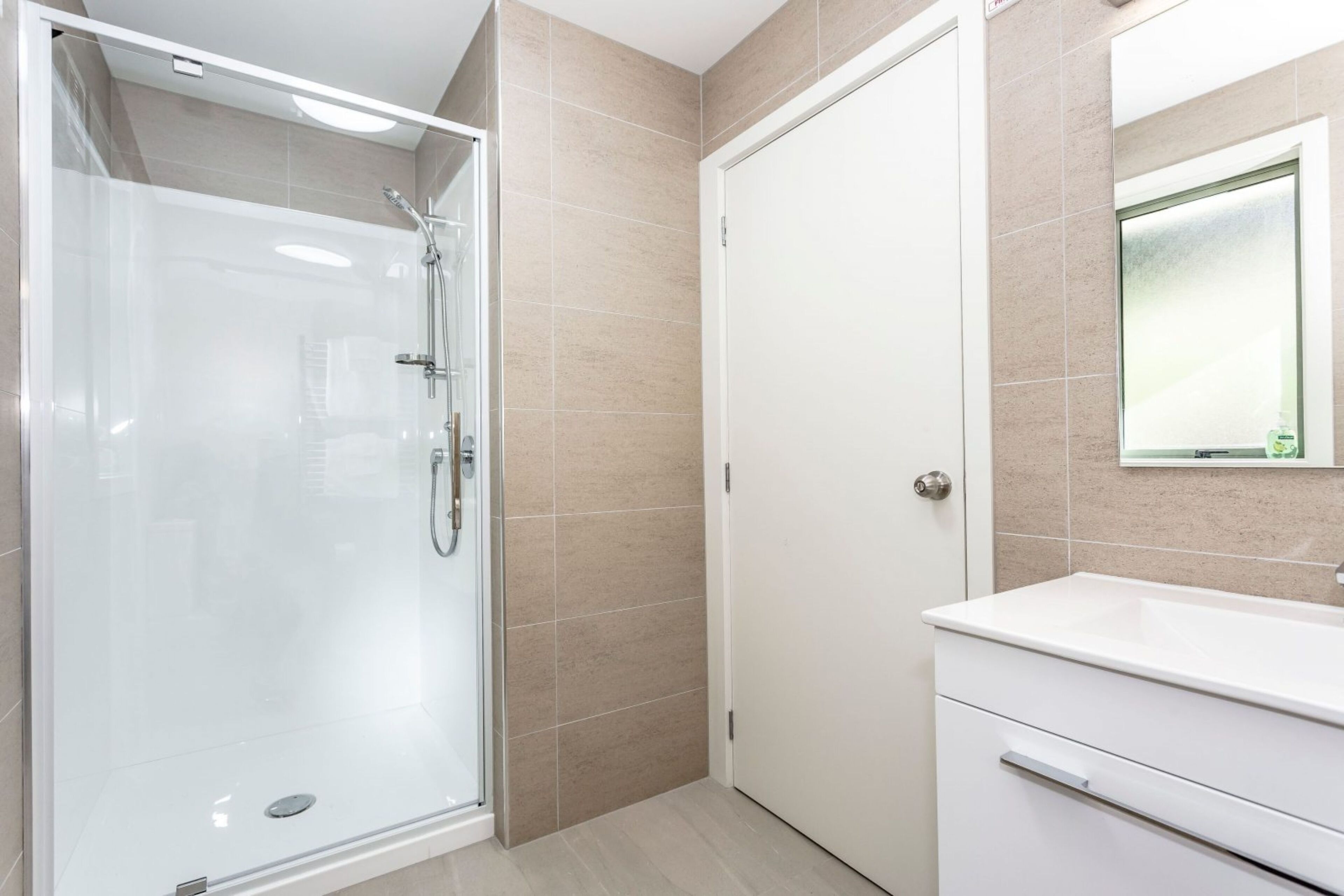 A modern bathroom with a glass-enclosed shower, a white vanity with a mirror, and beige tiled walls.