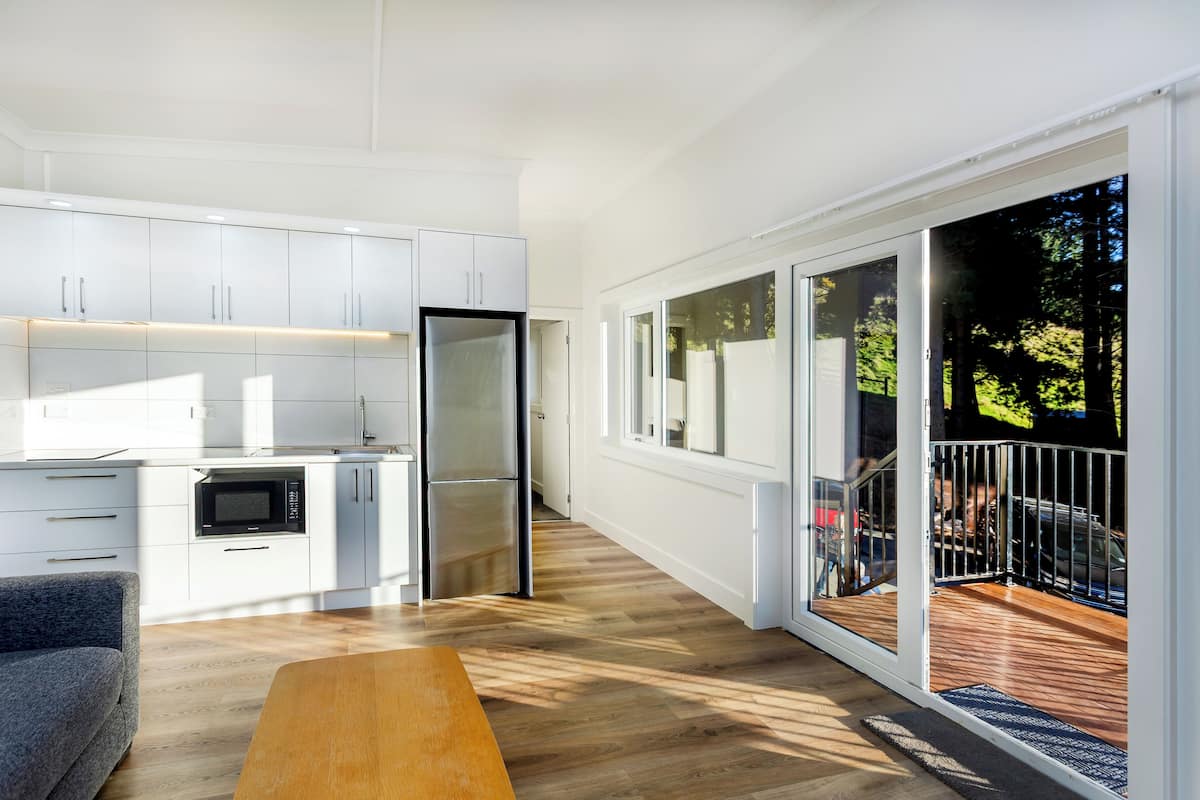 Una cocina moderna con armarios blancos, microondas integrado y refrigerador. En la zona de estar hay un banco de madera y una puerta corrediza de vidrio que da a una terraza exterior.