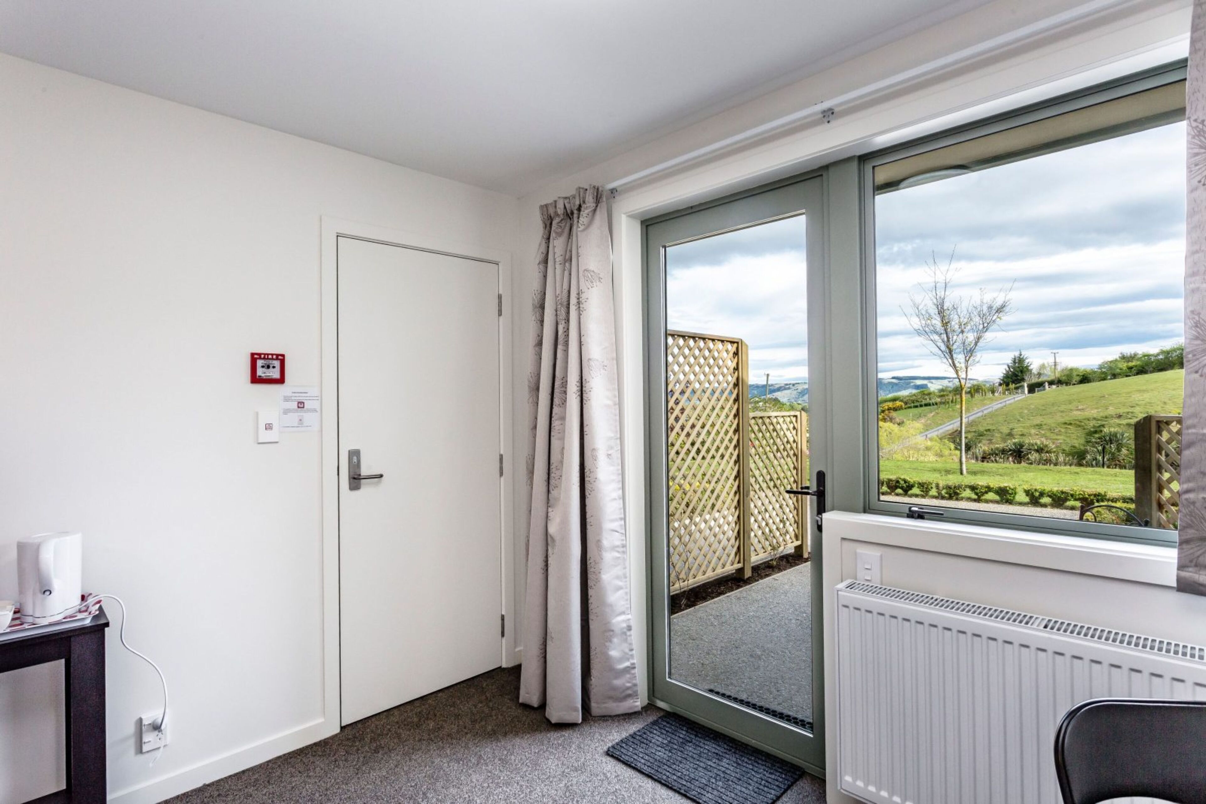 A room with a door, a radiator, a window with a view of greenery, and a small table with a tissue box.