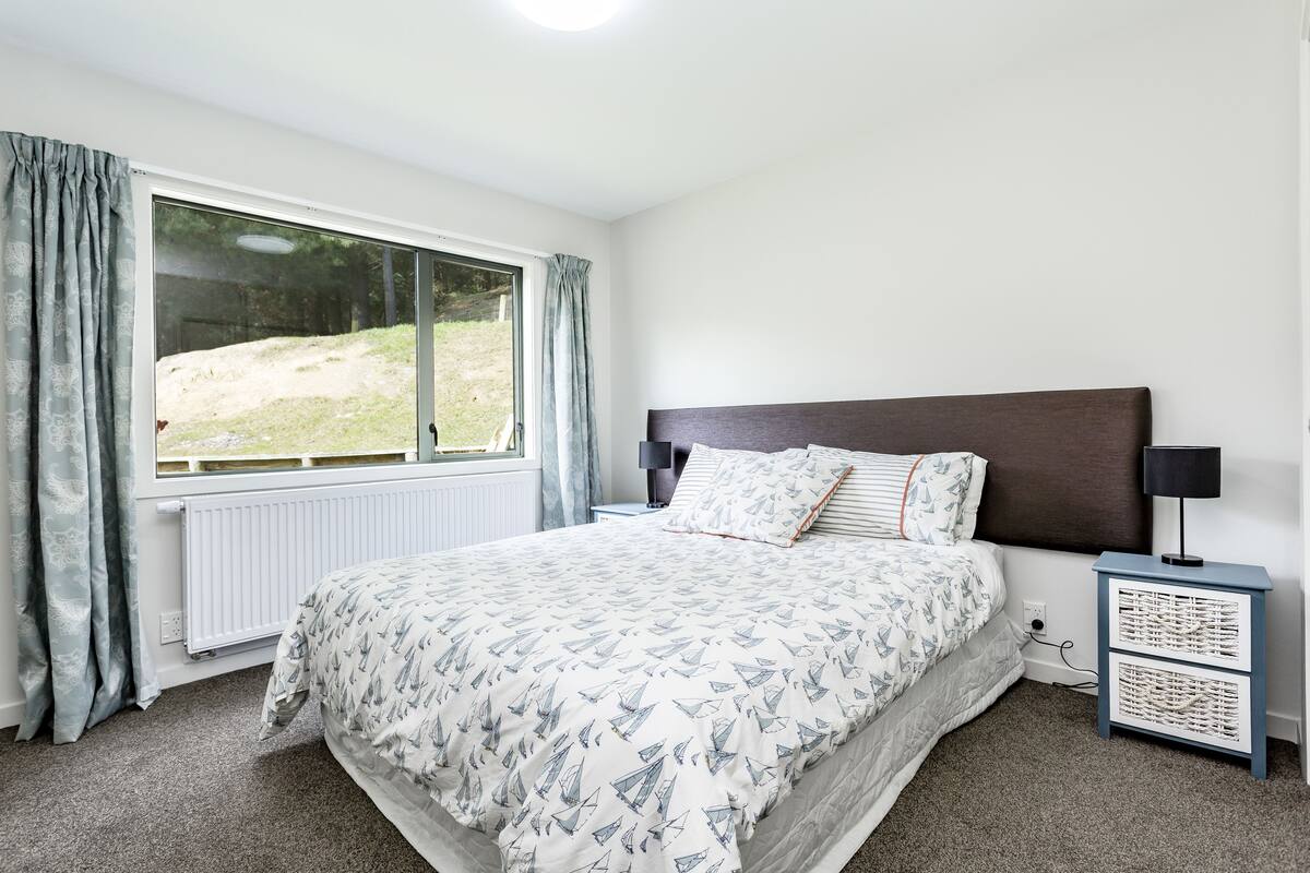 Un dormitorio con un ventanal grande, una cama con ropa de cama estampada, una mesita de noche con lámpara y vistas a un área verde exterior.