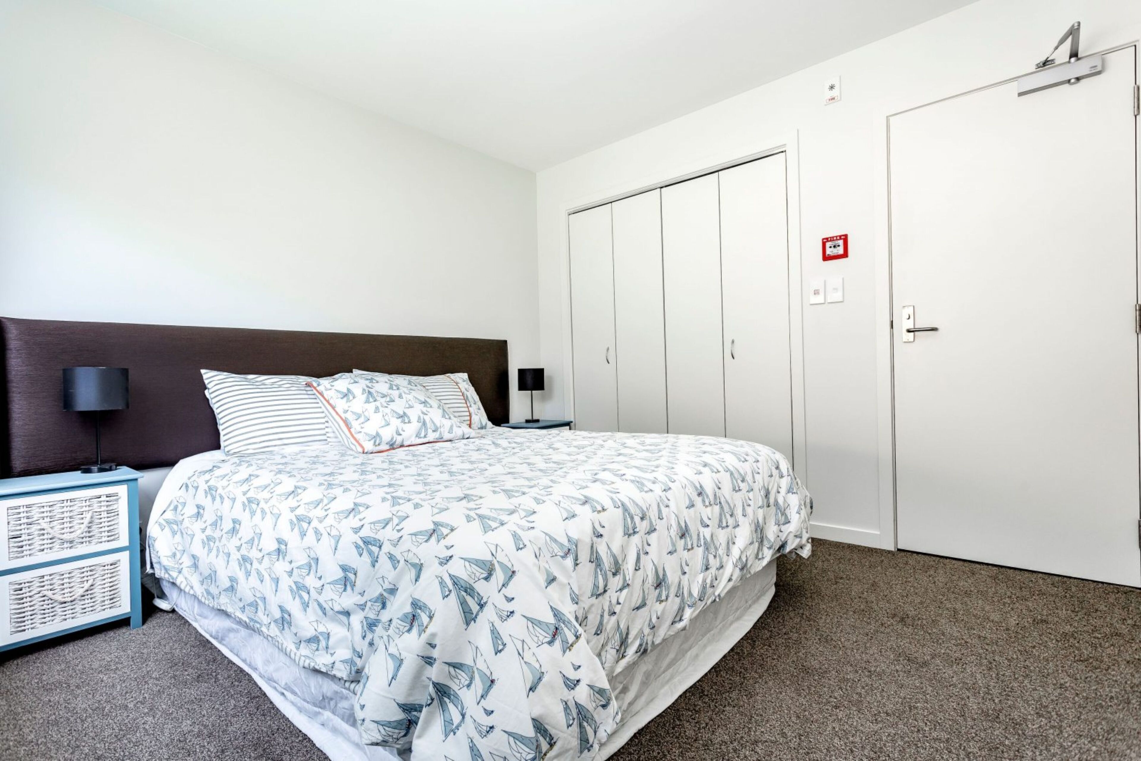 A bedroom with a bed, a nightstand, a wardrobe, and a door with a fire alarm.