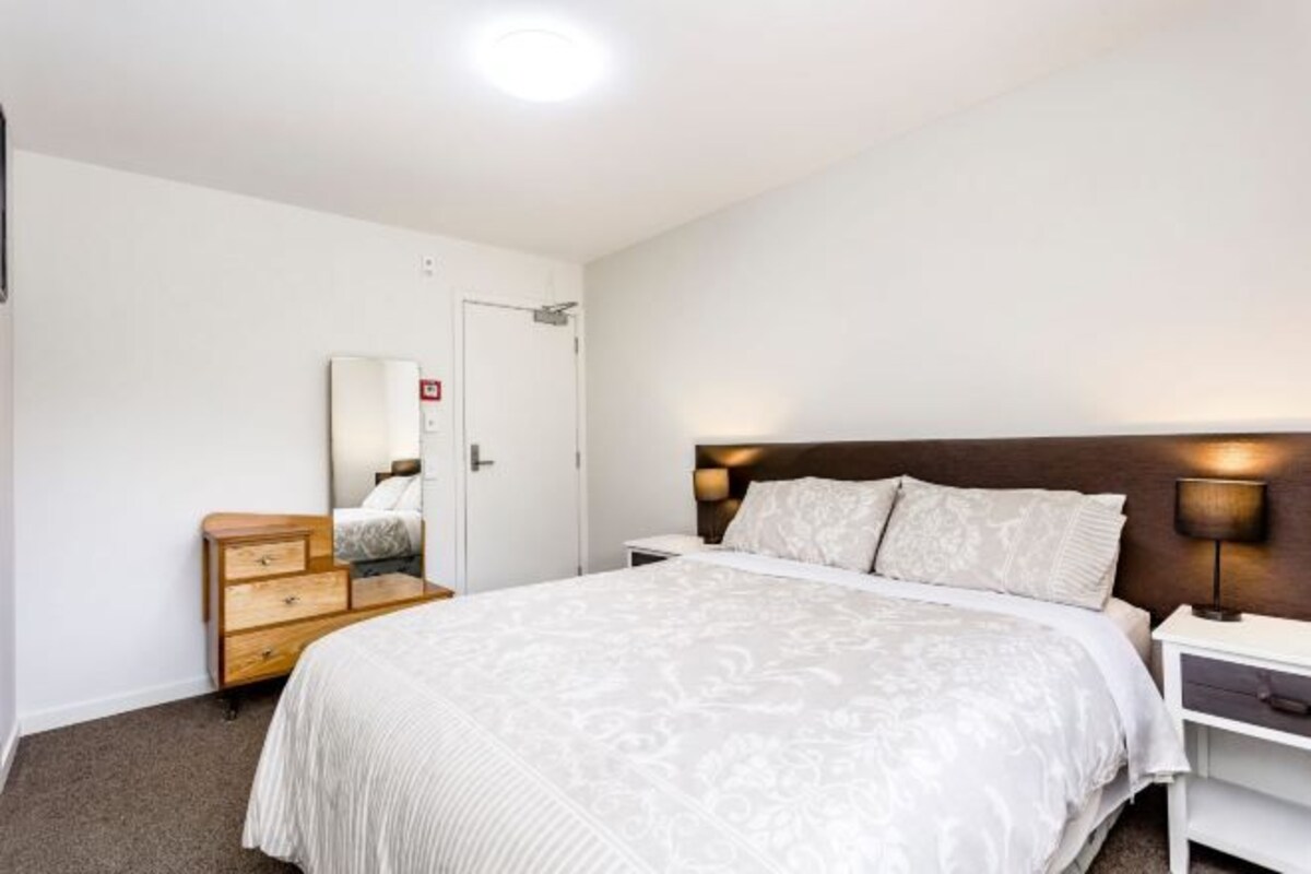 A hotel room with a bed, bedside table, lamp, dresser, and a door.