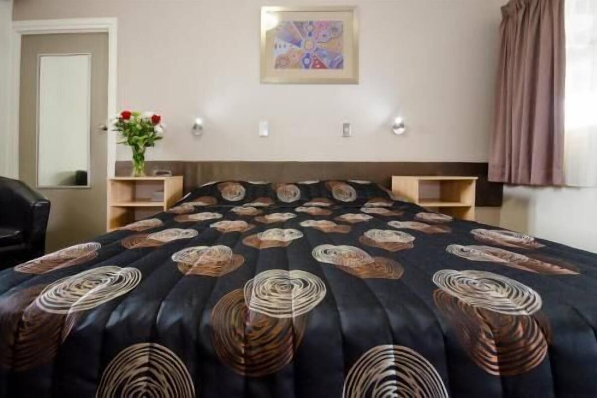 Une chambre d’hôtel avec un lit, un bureau, une chaise, un vase de fleurs, un tableau au mur et une fenêtre avec des rideaux.