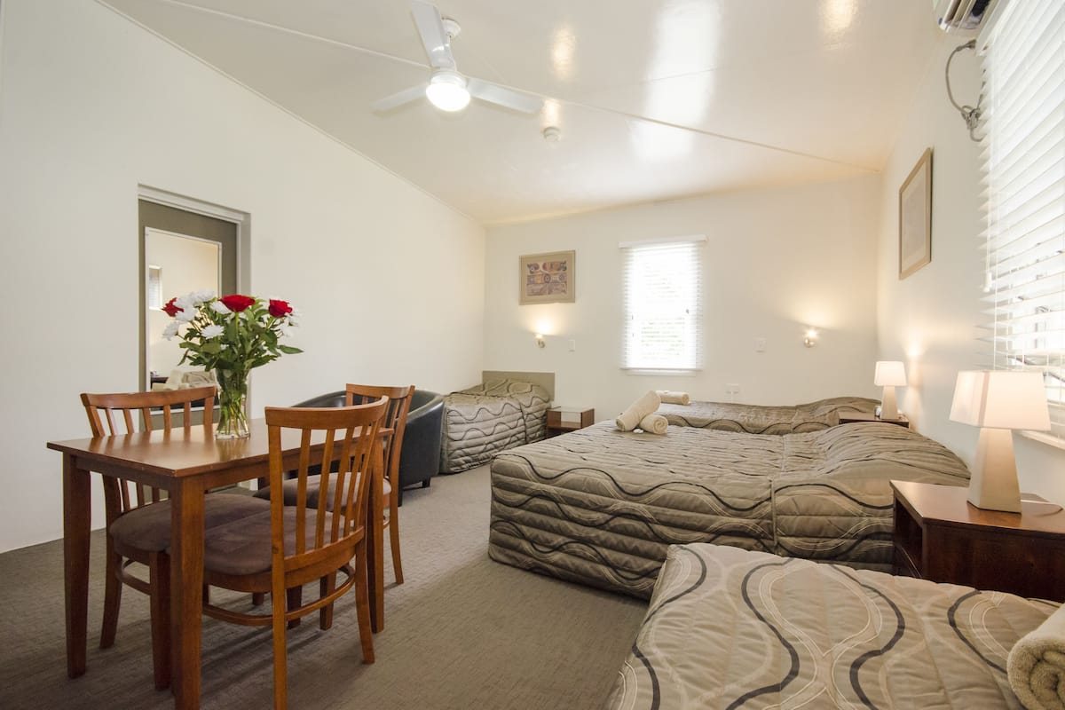 Une chambre avec deux lits, une table à manger avec des chaises, un vase avec des fleurs et un ventilateur de plafond.