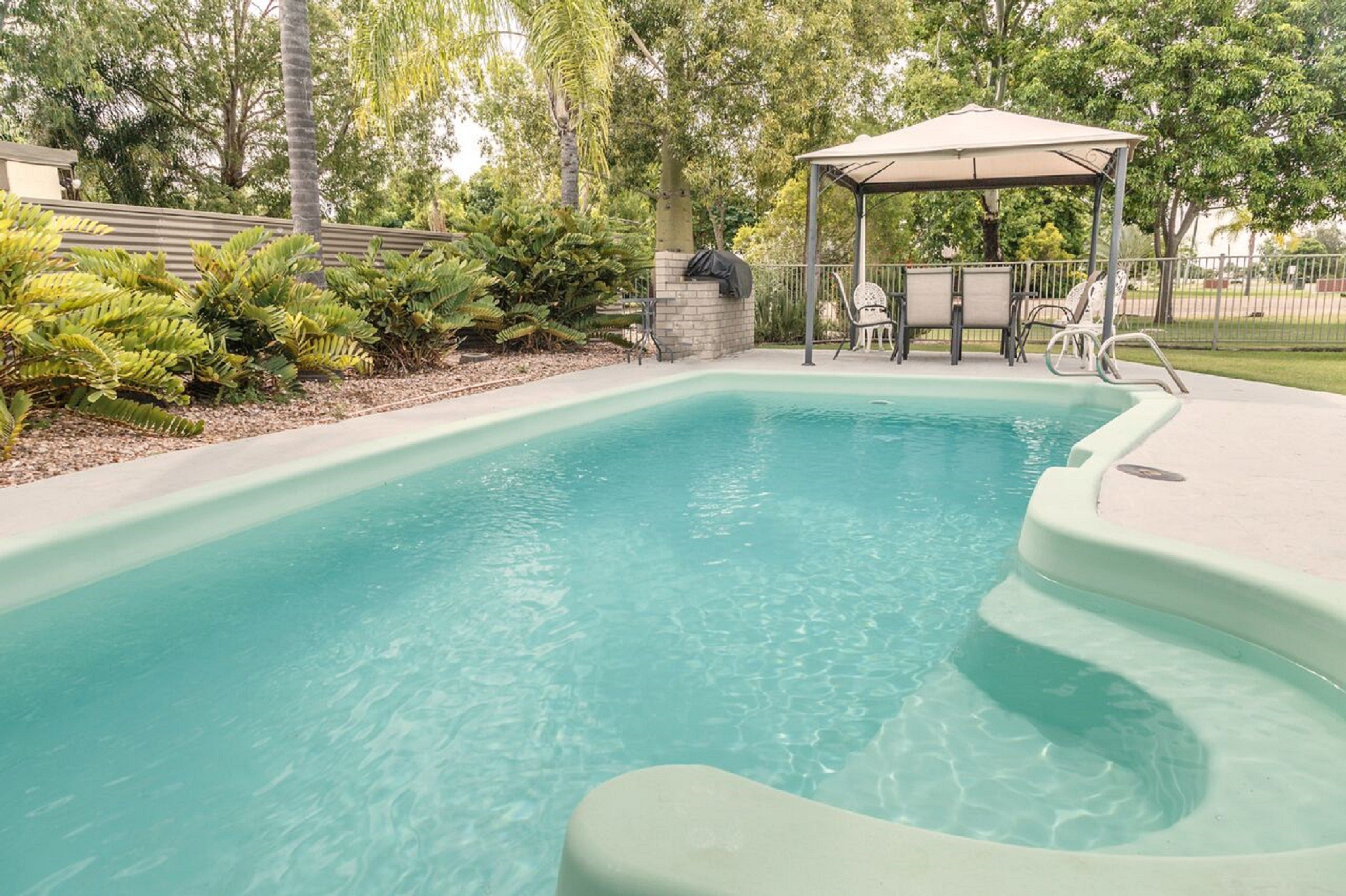 A rectangular pool with a curved edge, surrounded by a concrete deck. There is a covered seating area with chairs and a table, and a small structure with a roof.