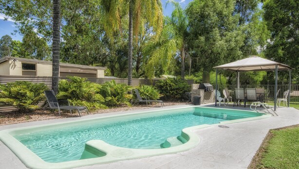 A swimming pool with a sunshade, surrounded by lush greenery and a small pavilion. - Biloela Countryman Motel (Biloela)
