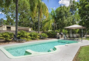 A swimming pool with a sunshade, surrounded by lush greenery and a small pavilion. - Biloela Countryman Motel (Biloela)