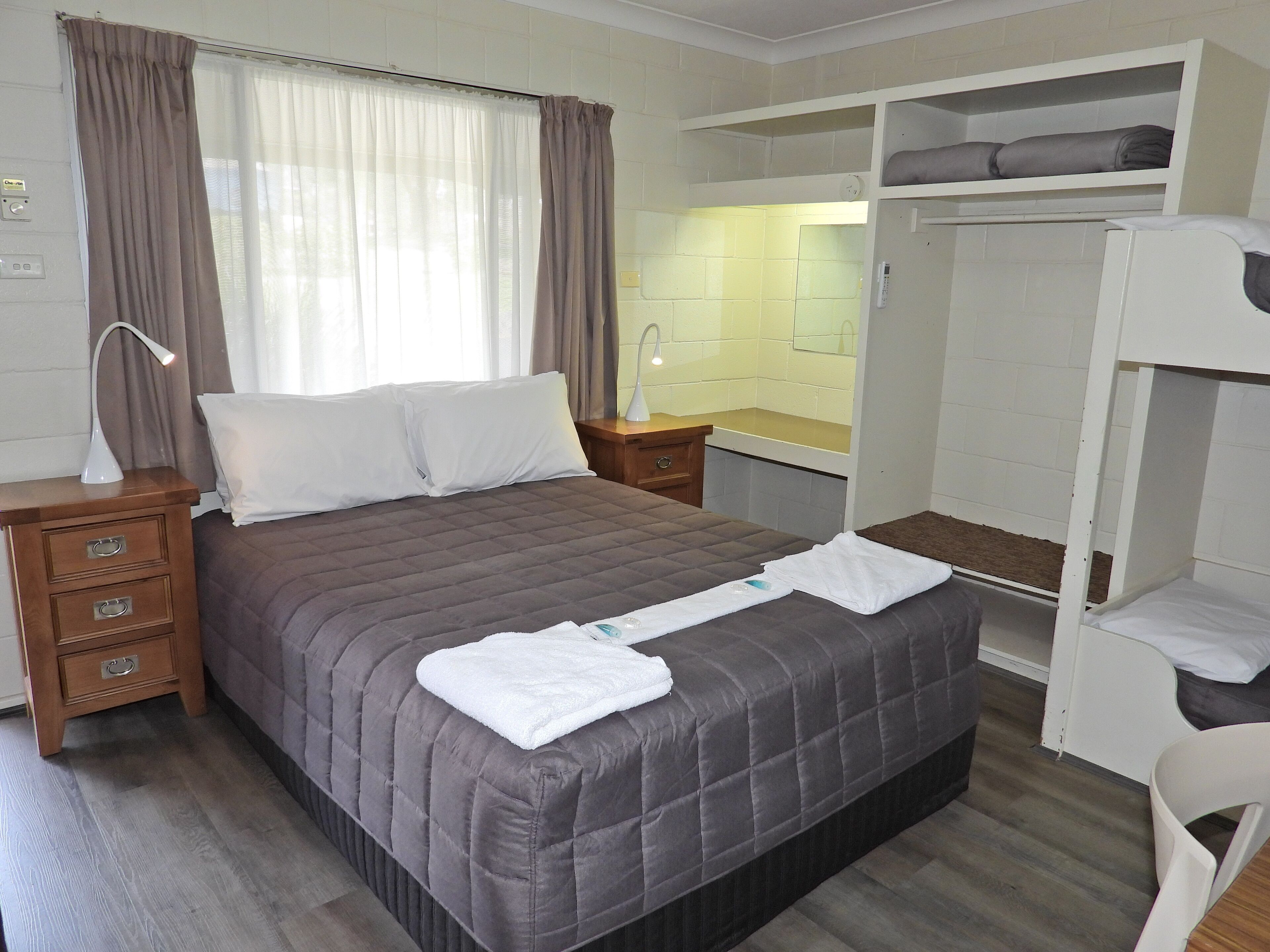 A hotel room with a bed, a bedside table with a lamp, a small desk with a mirror, a sink, and a wardrobe.