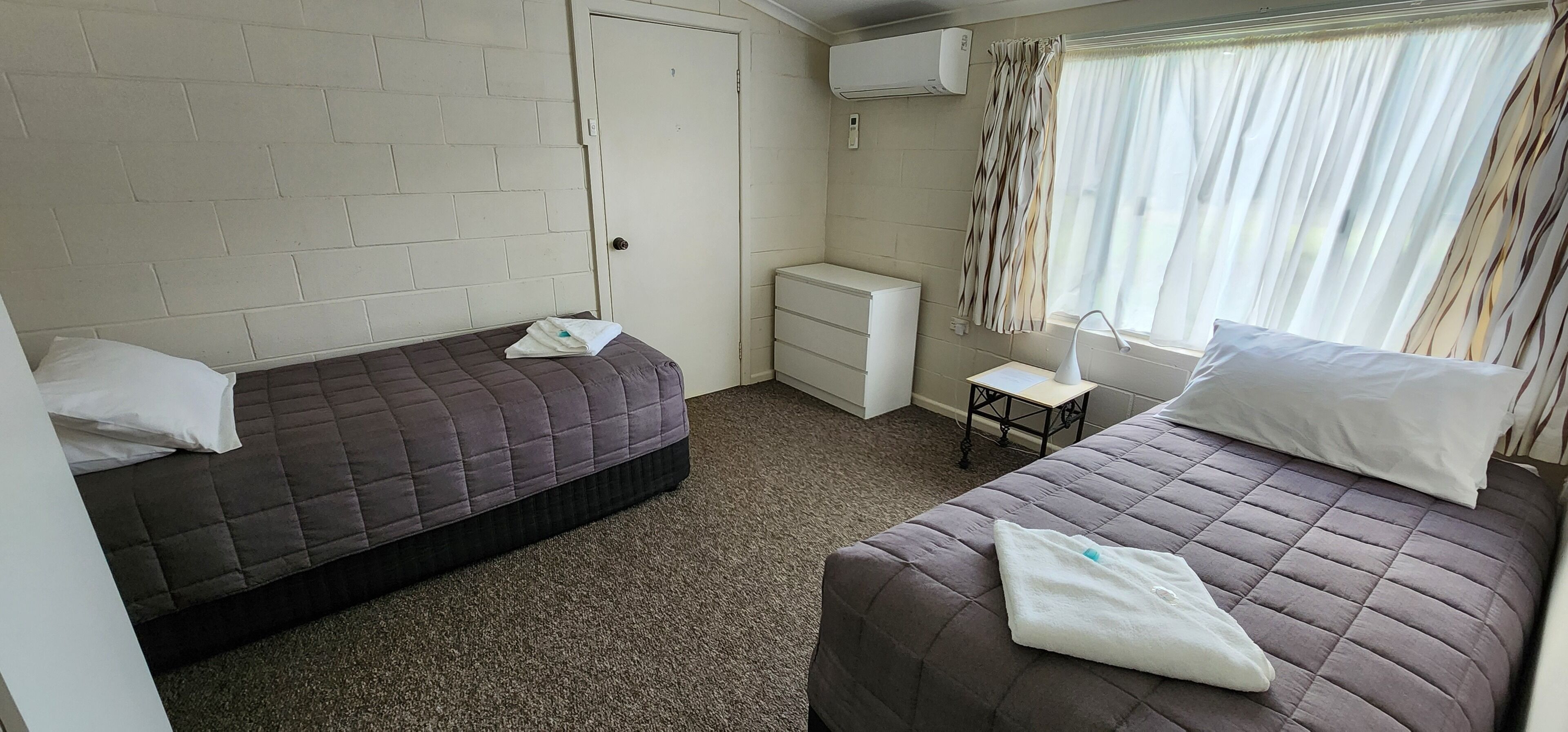 A room with two beds, a small table, a chair, a radiator, and a window with curtains.