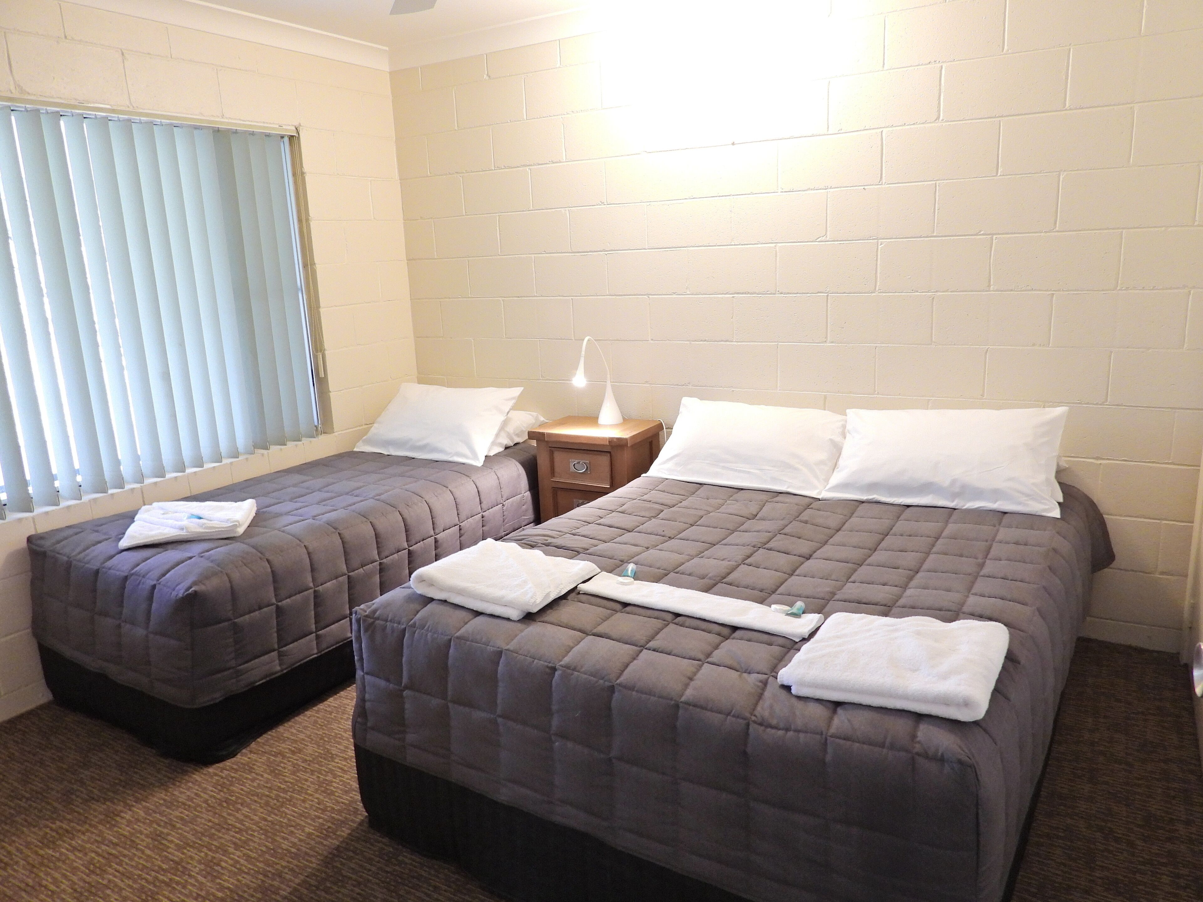 A hotel room with two beds, a nightstand with a lamp, and a window with blinds.