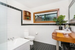Un baño con inodoro blanco, un lavabo blanco con encimera de madera, una bañera y una ventana con marco de madera.