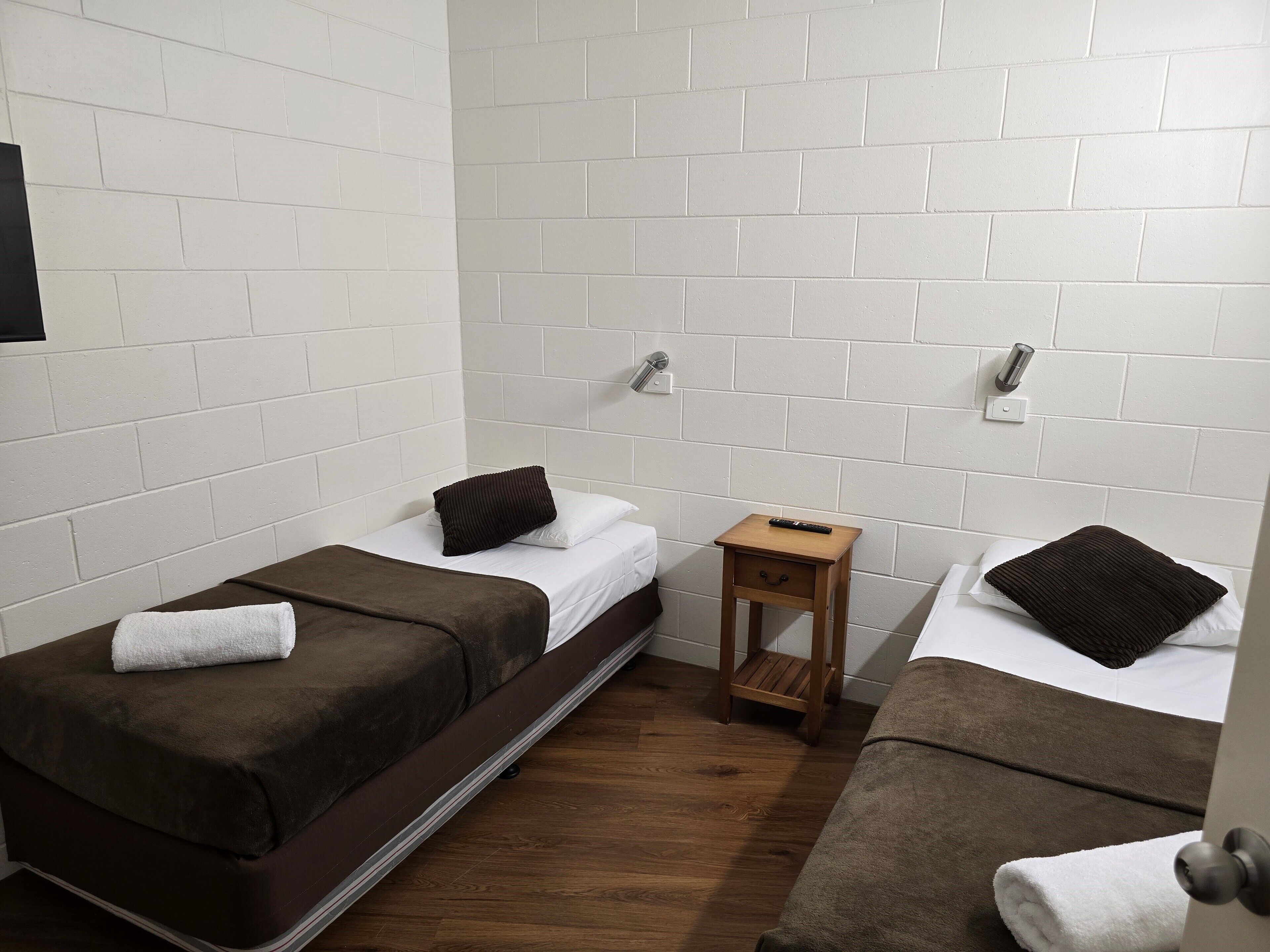 Een kleine, schone kamer met twee bedden, elk met een kussen en een dekbed. Er is een klein houten nachtkastje met een afstandsbediening erop.