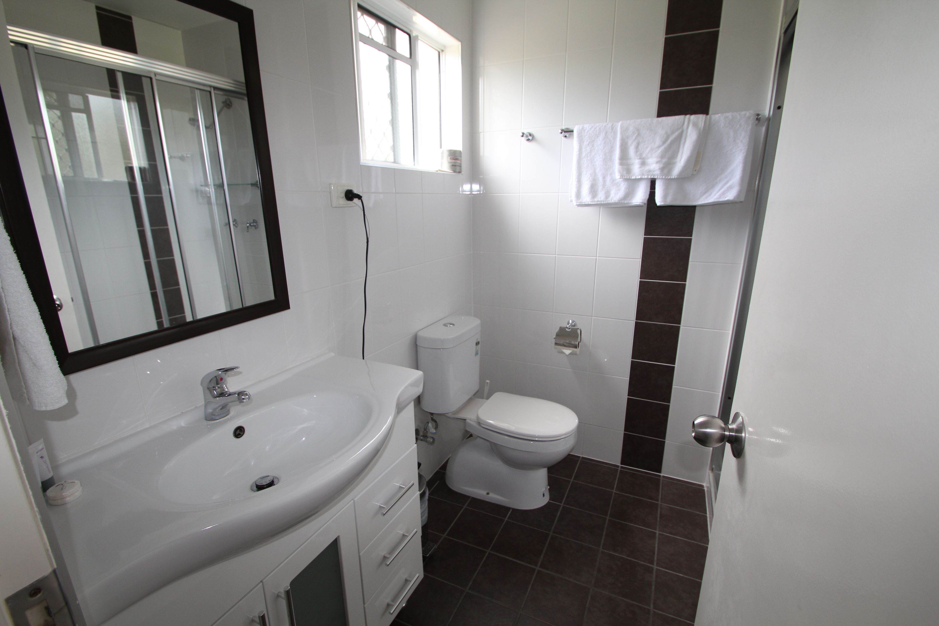 Un baño con lavamanos blanco, espejo e inodoro. Hay una ventana sobre el inodoro y una toalla colgada en la pared.