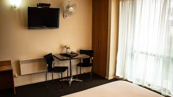 A hotel room with a bed, a desk with two chairs, a flat-screen TV, a standing fan, and a window with sheer curtains.