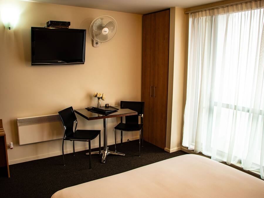 A hotel room with a bed, a desk with two chairs, a flat-screen TV, a standing fan, and a window with sheer curtains.