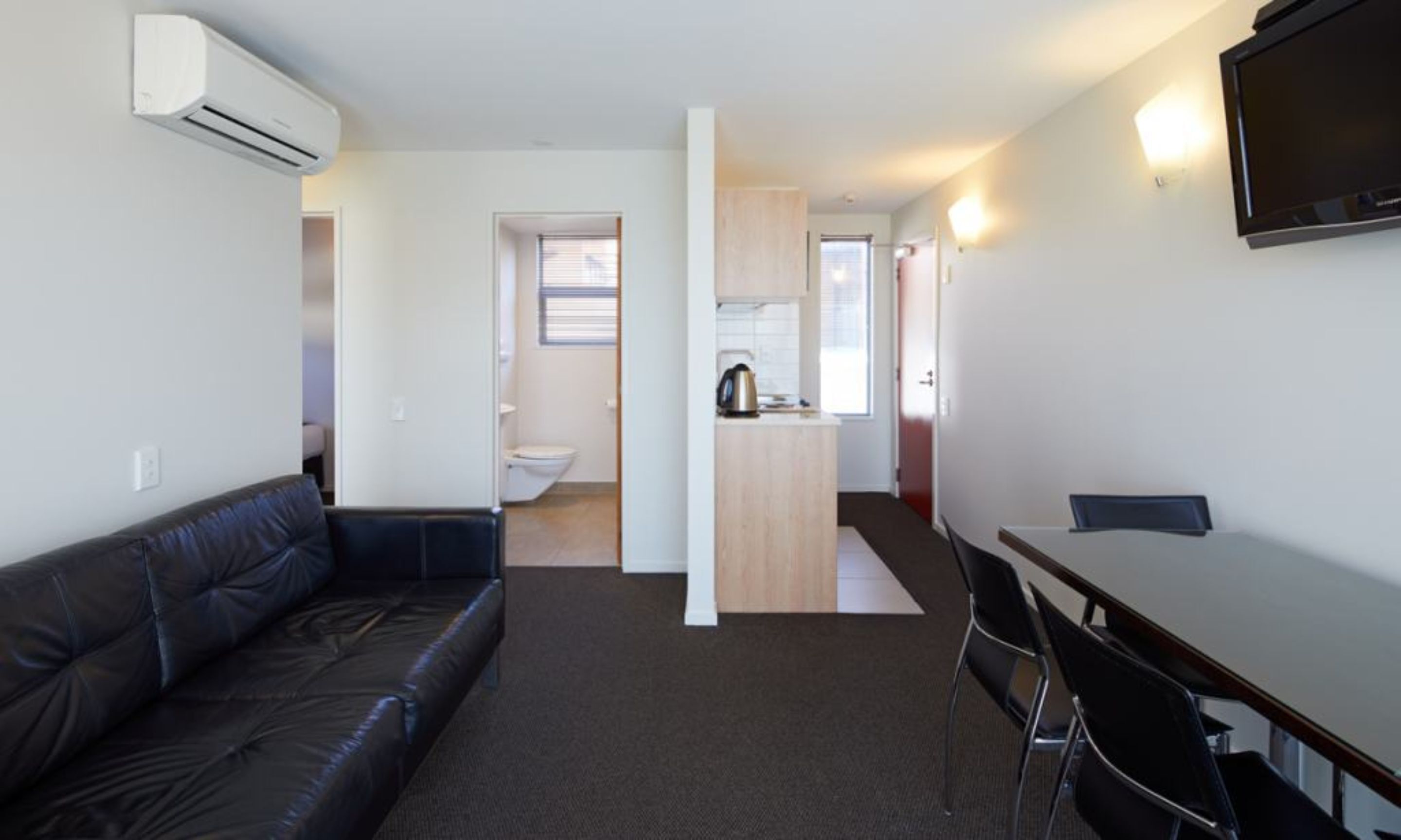 A compact living space with a sofa, a dining table with chairs, a kitchenette, and a TV mounted on the wall.