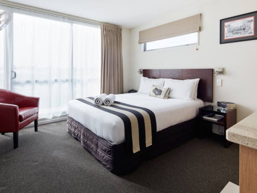 A hotel room with a large bed, a red armchair, a nightstand, and a window with curtains.
