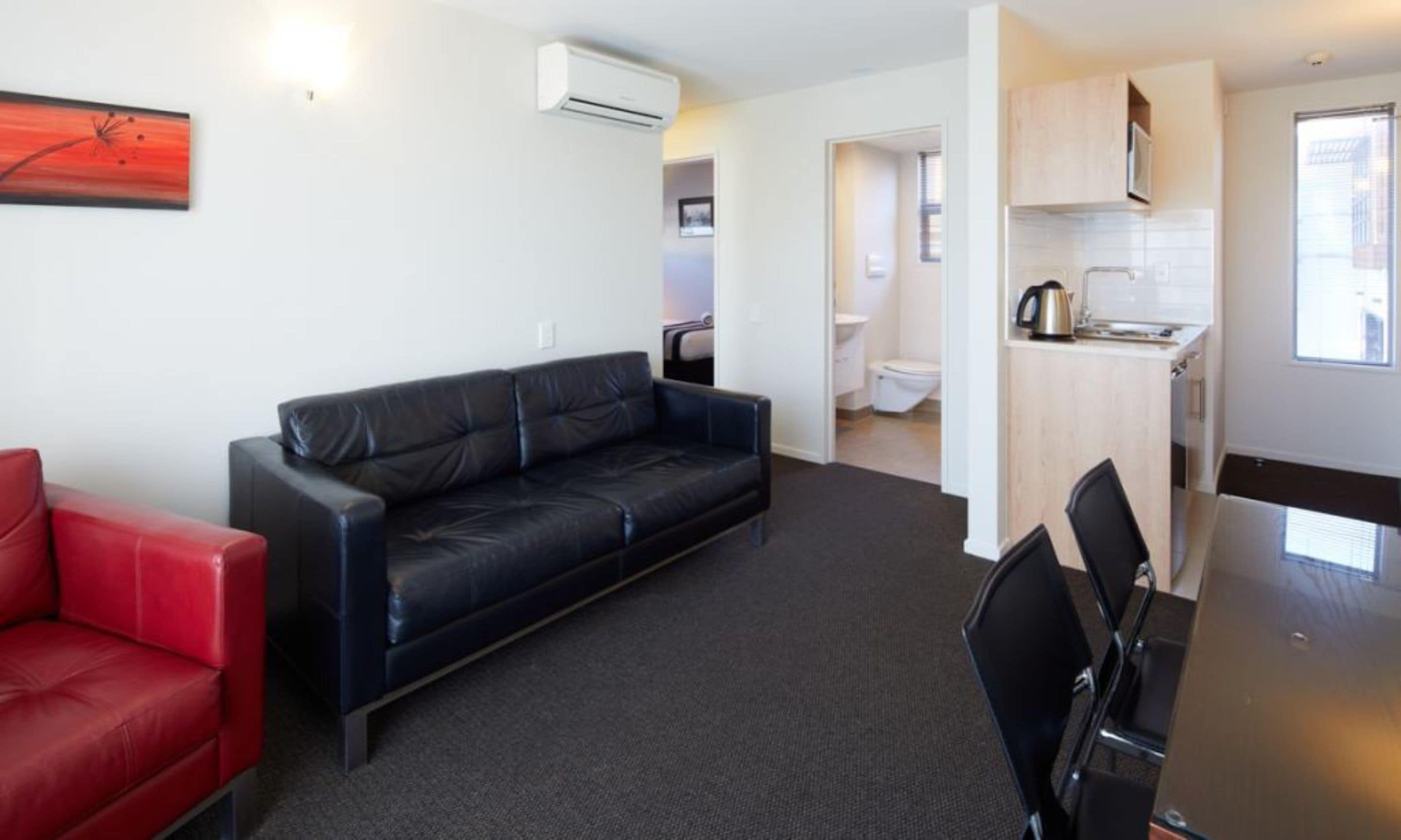 A modern living room with a black leather sofa, a red armchair, and a dining table with chairs. There is a kitchenette with a stove and a window.