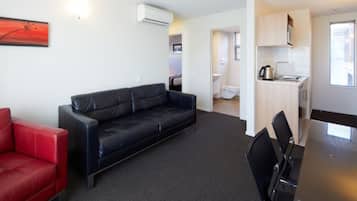 A modern living room with a black leather sofa, a red armchair, and a dining table with chairs. There is a kitchenette with a stove and a window.