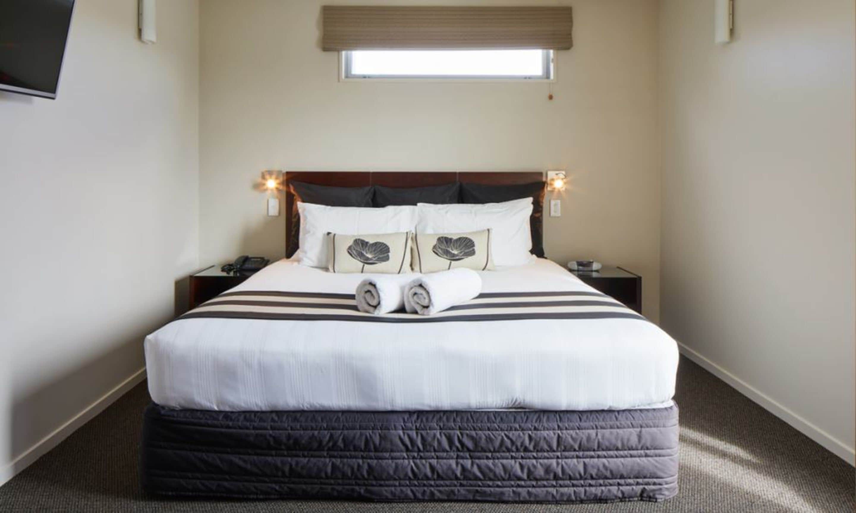 Una cama bien hecha con una colcha a rayas, dos almohadas con diseño de hojas y una toalla enrollada sobre la cama. Hay una mesita de noche con un teléfono y una lámpara a cada lado de la cama.