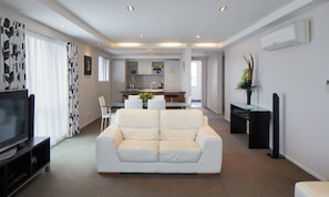 Uma sala de estar moderna com um sofá branco, uma TV de tela plana e um espaço de jantar com mesa e cadeiras de vidro.