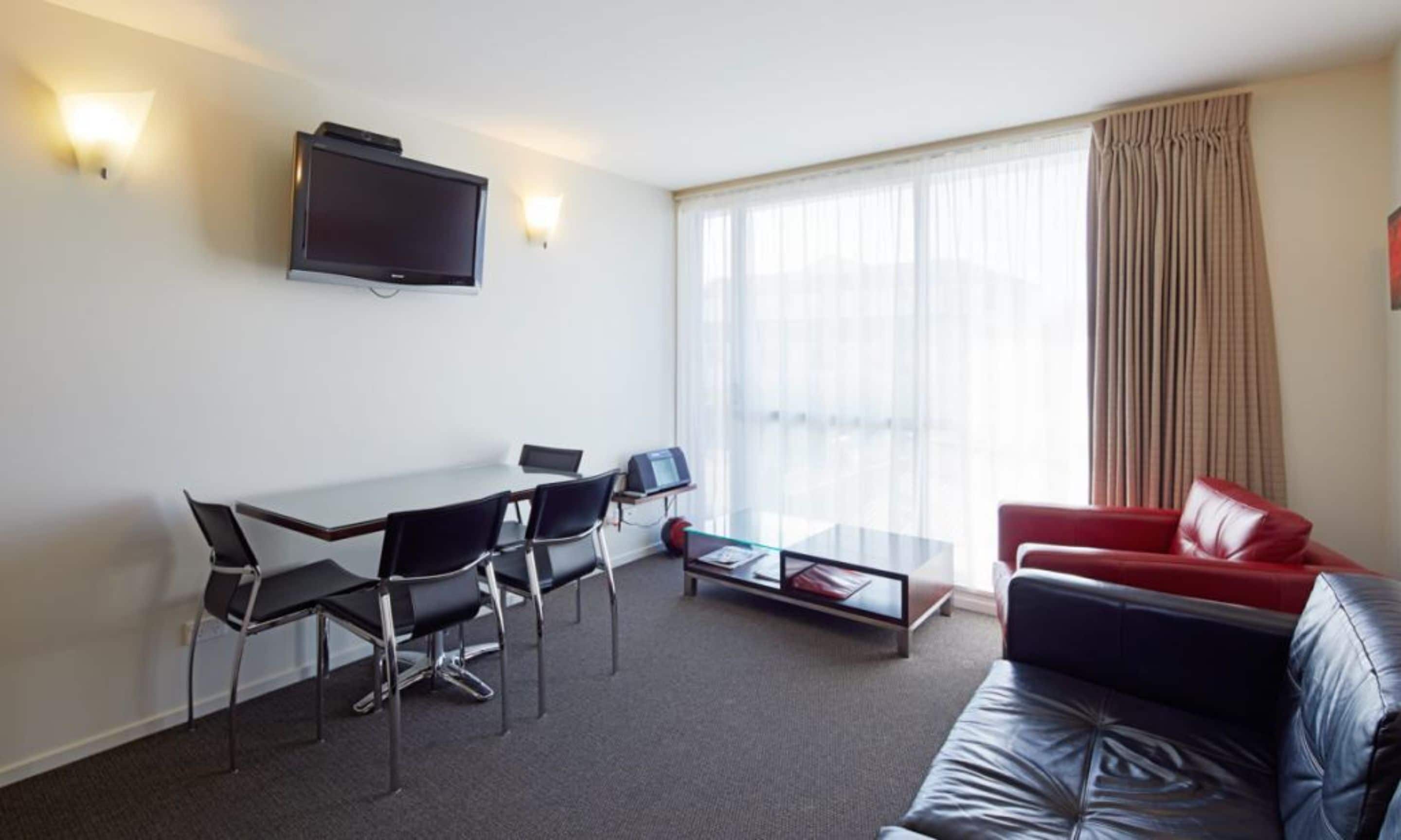 A modern hotel room with a flat-screen TV mounted on the wall, a dining area with a table and chairs, a red leather sofa, and a large window with curtains.