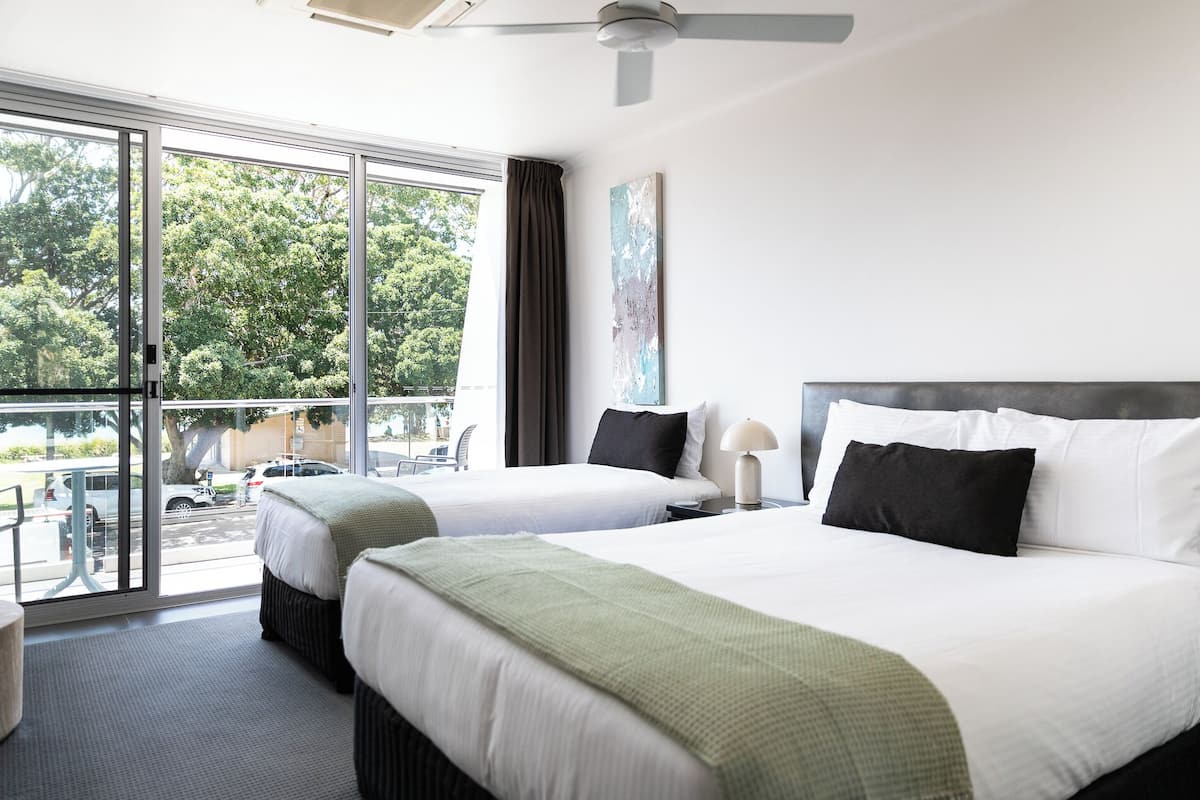 Ein Hotelzimmer mit zwei Betten, einem großen Fenster mit Blick auf Bäume, einem Deckenventilator und einem Gemälde an der Wand.
