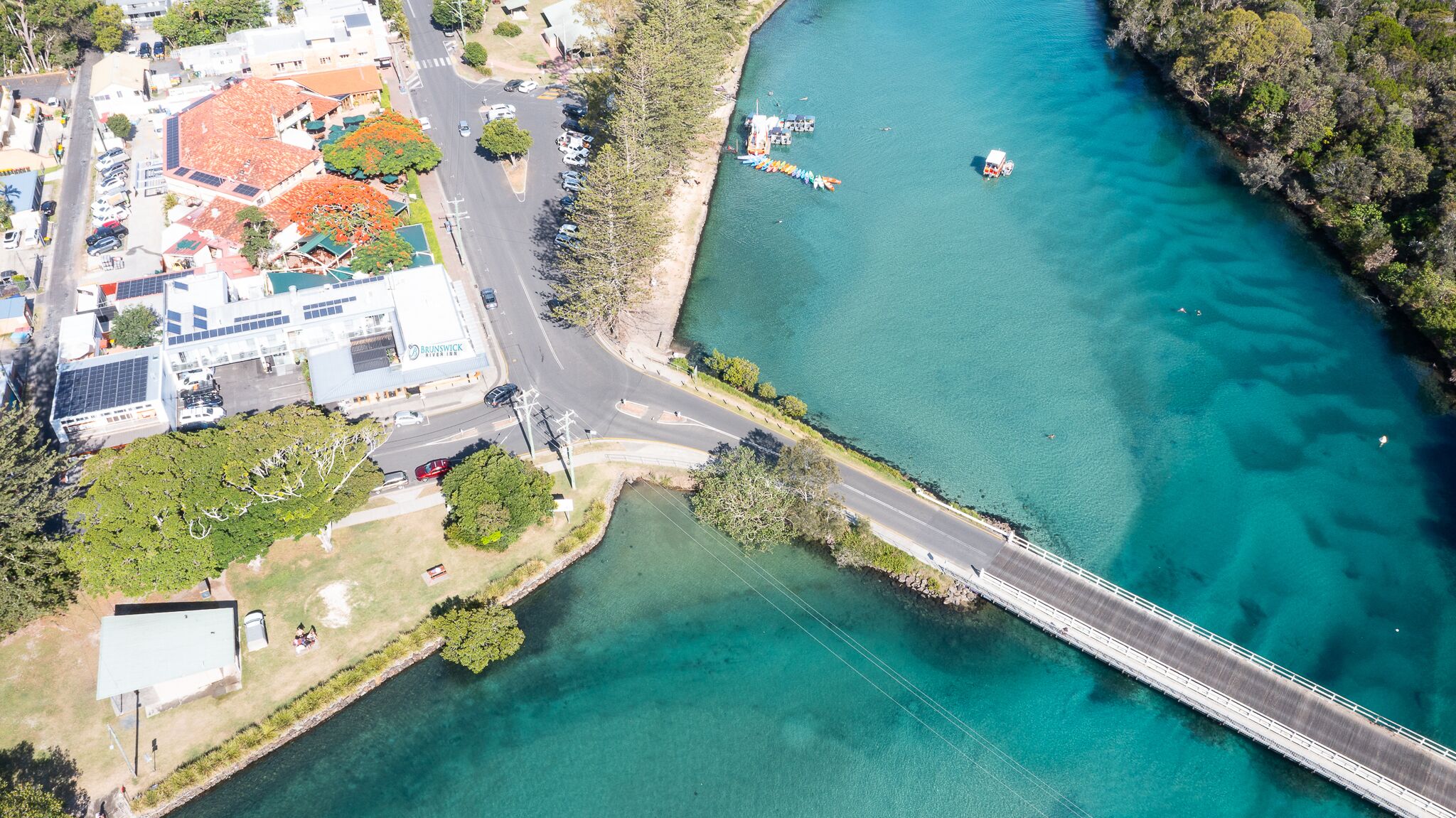 An aerial view of a coastal area with a bridge, clear turquoise water, and a building with solar panels.