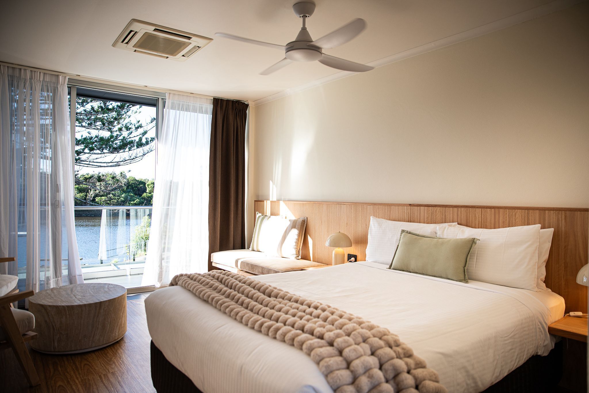 Habitación de hotel con cama, una mesita de madera redonda, ventilador de techo y vista a un cuerpo de agua a través de la ventana.