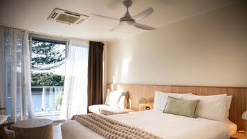 Habitación de hotel con cama, una mesita de madera redonda, ventilador de techo y vista a un cuerpo de agua a través de la ventana.