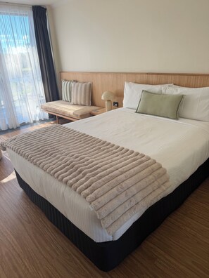 A hotel room with a large bed, a bedside table with a lamp, a bench with pillows, and a view through the window.