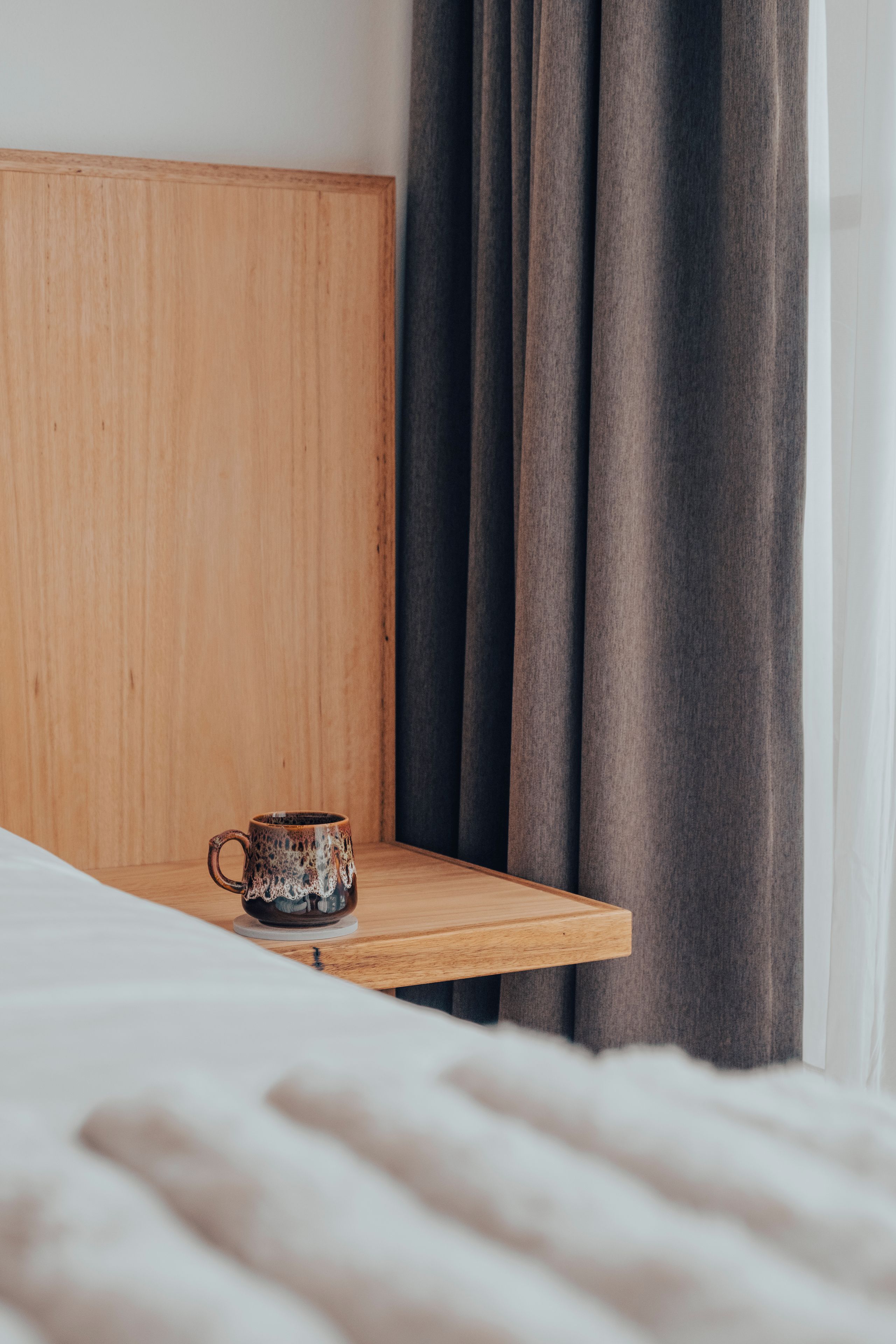 Una mesita de noche de madera con una taza encima, una cama con ropa blanca y una cortina oscura al fondo.