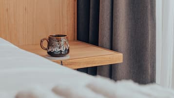 Una mesita de noche de madera con una taza encima, una cama con ropa blanca y una cortina oscura al fondo.