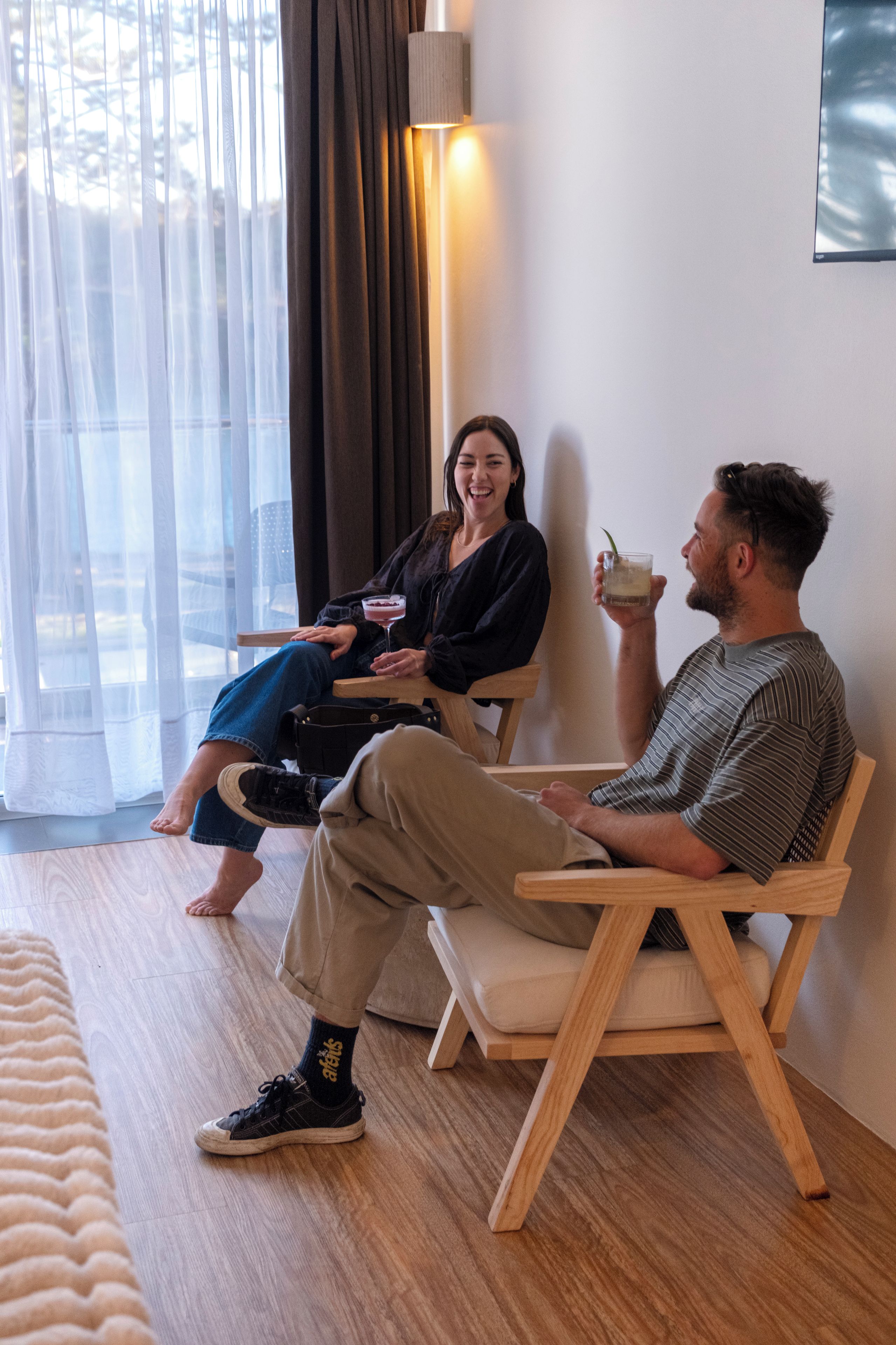 A room with a wooden floor, a sofa, a wooden chair, and a person holding a drink.