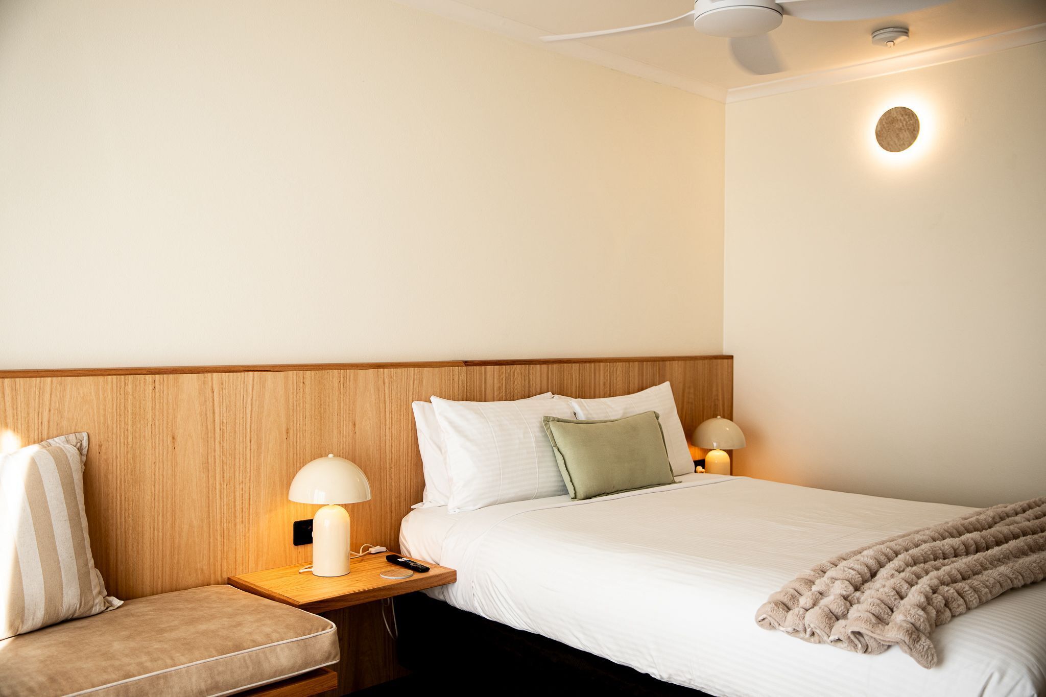 Habitación de hotel con cabecera de madera, cama con ropa blanca, una manta color beige, una mesita de noche con lámpara y una lámpara de pared.