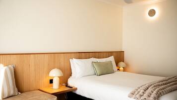 Habitación de hotel con cabecera de madera, cama con ropa blanca, una manta color beige, una mesita de noche con lámpara y una lámpara de pared.