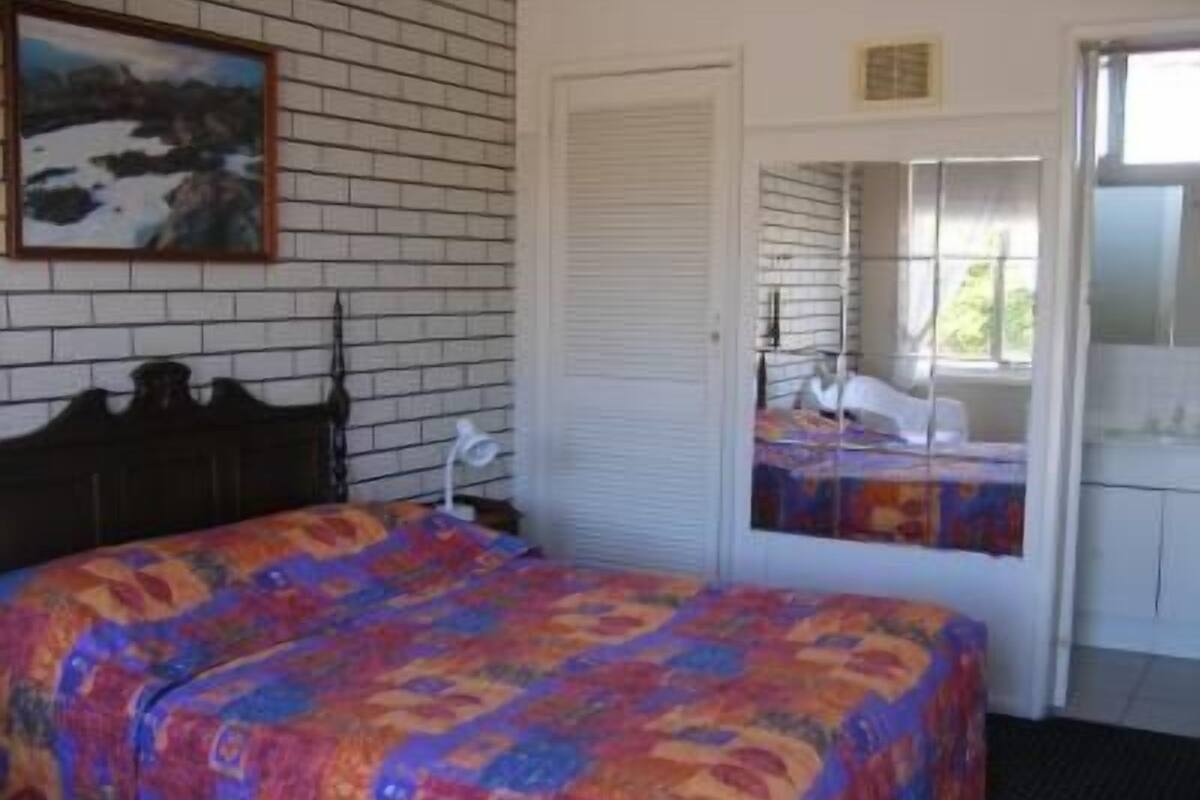 Un dormitorio con cama, mesita de noche, lámpara, ventana con persianas y un cuadro en la pared.
