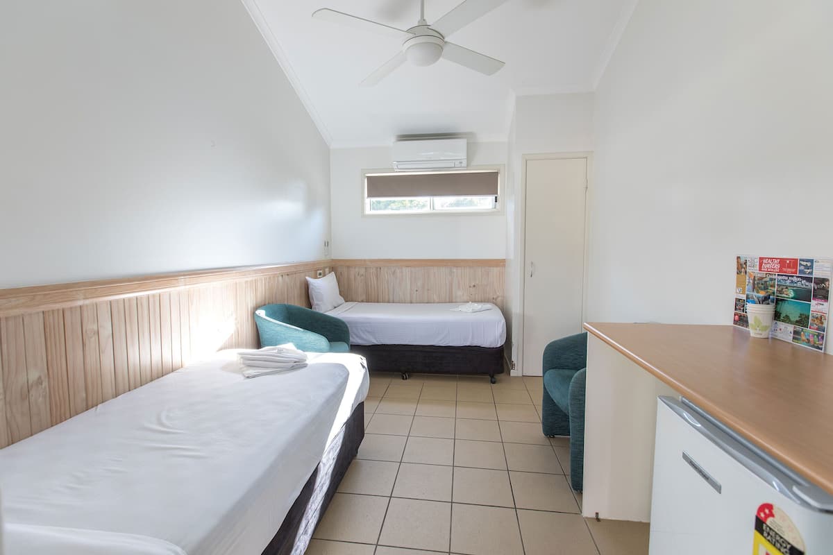 Une petite chambre propre, avec deux lits simples, une tête de lit en bois, un ventilateur de plafond, une petite kitchenette et une fenêtre avec des stores.